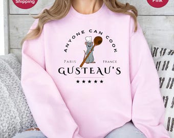 May include: A light pink sweatshirt with the text "Anyone Can Cook" in a circle around a cartoon rat wearing a chef's hat and holding a wooden spoon. The text "Paris" and "France" are below the circle, and the text "Gusteau's" is below that, with five stars underneath.
