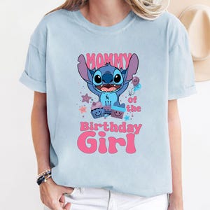 K&ouml;nnte beinhalten: Hellblaues T-Shirt mit einer blauen Stitch-Figur aus Lilo & Stitch. Das Shirt tr&auml;gt den Schriftzug "Mommy of the Birthday Girl" in rosa.