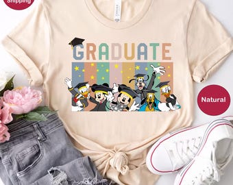 May include: A light beige t-shirt featuring a retro-style graphic of Disney characters wearing graduation caps and the word "GRADUATE" in colourful letters. The characters include Mickey Mouse, Minnie Mouse, Goofy, Donald Duck, and Pluto.