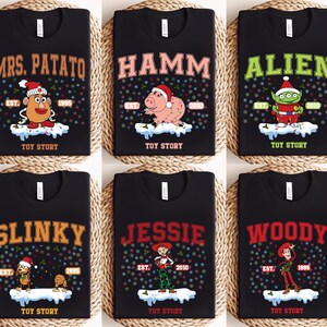May include: Six black t-shirts with different Toy Story characters printed on them. Each shirt has a festive Christmas theme with a snowy background and colorful lights. The characters include Mrs. Potato Head, Hamm, Alien, Slinky Dog, Jessie, and Woody.