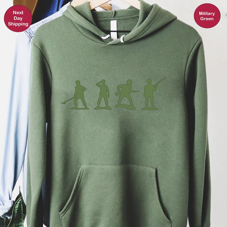 Puede incluir: Una sudadera con capucha verde con un gr&aacute;fico de cinco soldados de juguete en fila. Los soldados est&aacute;n todos mirando en la misma direcci&oacute;n y sostienen diferentes armas. La sudadera es de color verde militar.