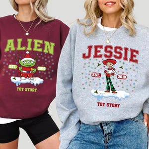 May include: Two Christmas sweaters with Toy Story characters. The red sweater features the alien with the text "Alien Est. 2010 Toy Story". The gray sweater features Jessie with the text "Jessie Est. 2010 Toy Story".