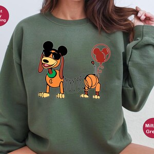May include: A green sweatshirt with a cartoon image of a dog with a spring attached to its tail. The dog is wearing a red bow tie and a red balloon with Mickey Mouse ears is attached to the spring. The text "Next Day Shipping" is in a red circle in the top left corner of the image.