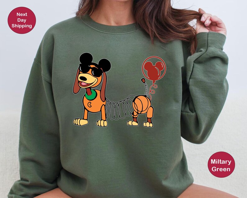 May include: A green sweatshirt with a cartoon image of a dog with a spring attached to its tail. The dog is wearing a red bow tie and a red balloon with Mickey Mouse ears is attached to the spring. The text "Next Day Shipping" is in a red circle in the top left corner of the image.