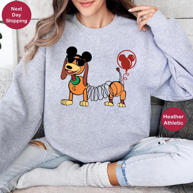May include: A gray sweatshirt with a cartoon image of a dog with a spring body and Mickey Mouse ears and sunglasses. The dog is holding a red balloon with a Mickey Mouse head on it. The text "Heather Athletic" is printed on the bottom right corner of the sweatshirt.