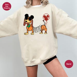 May include: A sand-colored sweatshirt with a cartoon image of a dog with a springy tail and Mickey Mouse ears. The dog is wearing sunglasses and has a red balloon with Mickey Mouse ears attached to its tail.