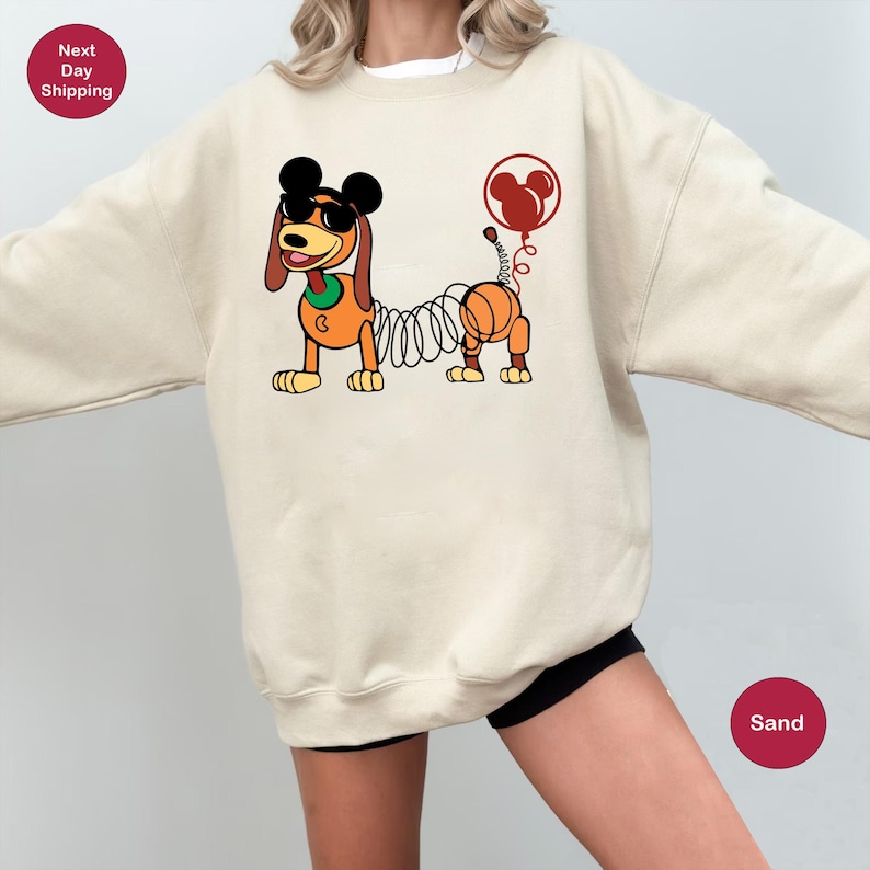 May include: A sand-colored sweatshirt with a cartoon image of a dog with a springy tail and Mickey Mouse ears. The dog is wearing sunglasses and has a red balloon with Mickey Mouse ears attached to its tail.