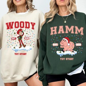 May include: Two cream and green Christmas themed sweatshirts with Toy Story characters. The cream sweatshirt features Woody with the text "WOODY EST. 1995 TOY STORY". The green sweatshirt features Hamm with the text "HAMM EST. 2010 TOY STORY".