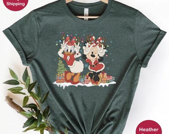 May include: A green heather forest t-shirt featuring a Christmas-themed design with Mickey Mouse and Donald Duck wearing Santa hats and holding presents. The design includes a Christmas tree and presents.