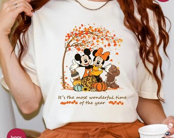 May include: White t-shirt with a graphic of Mickey Mouse and Minnie Mouse in fall attire. The graphic includes a tree with orange and yellow leaves, pumpkins, and a coffee cup. The text "It's the most wonderful time of the year" is printed below the graphic.