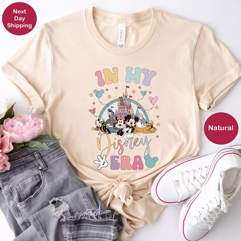May include: A beige t-shirt with a graphic print of Mickey Mouse and other Disney characters. The graphic includes the text "In My Disney Era" with a rainbow-colored background.