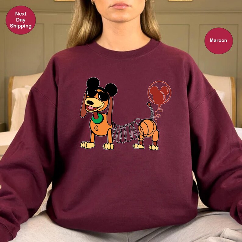 May include: A maroon sweatshirt with a cartoon image of a dog with a spring body and Mickey Mouse ears. The dog is wearing sunglasses and has a red balloon attached to its tail. The text "Maroon" is visible in the upper right corner of the image.
