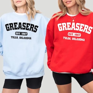 May include: Two women wearing sweatshirts with the text "Greasers Est. 1967 Tulsa, Oklahoma" in black and white lettering. One sweatshirt is light blue and the other is red.