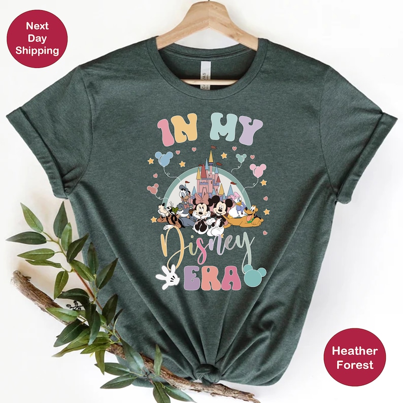 May include: A green t-shirt with the text "In My Disney Era" in rainbow colors. The design features a cartoon image of Mickey Mouse and other Disney characters in front of Cinderella's castle.