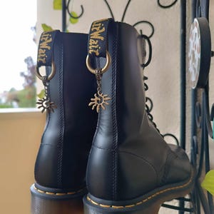 Puede incluir: Botas de cuero negro con costuras amarillas y tiradores. Cada bota tiene un charm de sol dorado colgando de un anillo. Las botas se exhiben en un estante de metal negro.