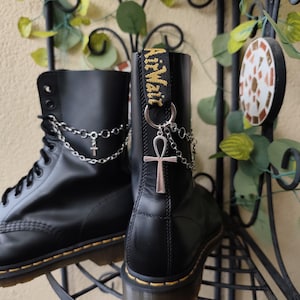 May include: Black leather boots with silver chain and Ankh charms. The boots have yellow stitching and the words "Air Wair" on the pull tab. The boots are displayed on a black metal shelf with decorative leaves.