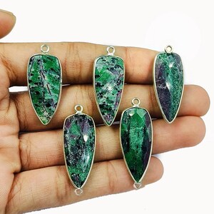 May include: Five gemstone pendants with a silver-colored bezel. The gemstones are a mix of green and dark red. Each pendant has a loop at the top for hanging. The pendants are teardrop-shaped.