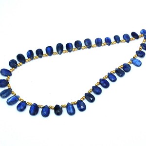 Peut inclure: Un collier composé de perles bleu foncé en forme de goutte d'eau, reliées par des entretoises dorées. Les perles ont un aspect poli et brillant et sont disposées de manière symétrique. Le fond est blanc.