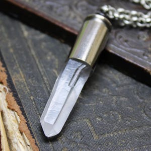 May include: A silver bullet casing pendant with a clear quartz crystal point attached. The pendant is hanging from a silver chain.