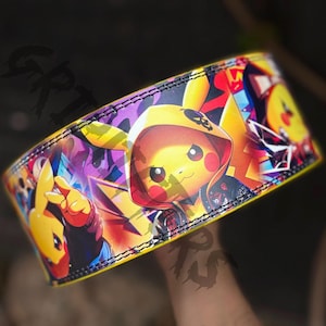 May include: A yellow and black leather wristband with a colorful graphic print of a cartoon character wearing a hooded jacket. The character has a skull and crossbones on the hood and is surrounded by a purple and yellow background.