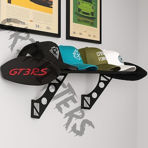 May include: A black wall shelf with a spoiler design holds five baseball caps. The caps are black, teal, olive green, and white, each with a different logo. A black sign with "GT3RS" in red lettering is also on the shelf. Two framed posters are above the shelf.
