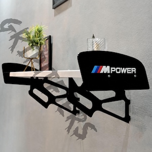 May include: A wall-mounted shelf featuring a white horizontal surface supported by black metal brackets. The side panels are black with the M POWER logo in blue, red, and white. The design mimics a car spoiler with a sleek, automotive-inspired aesthetic.