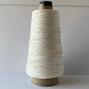 May include: A spool of natural white cotton twine wound on a cardboard cone. The twine is tightly wound and has a smooth, even texture.