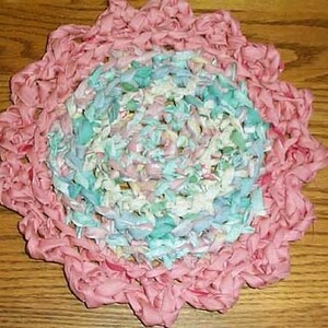 Puede incluir: Una alfombra redonda de crochet con un borde exterior rosa y un centro multicolor con tonos de azul, verde y blanco.