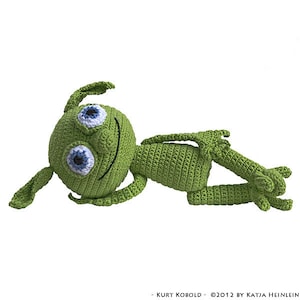 May include: A handmade green crochet creature with large blue and white eyes, lying on its side. The creature has a smiling face and outstretched arms and legs. The text "- KURT KOBOLD ©2012 BY KATJA HEINLEIN" is at the bottom.