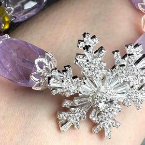 May include: A beaded bracelet featuring large, faceted amethyst stones and a sparkling, silver-toned snowflake pendant. The bracelet has a mix of purple, yellow, and clear beads. The snowflake is encrusted with clear crystals.