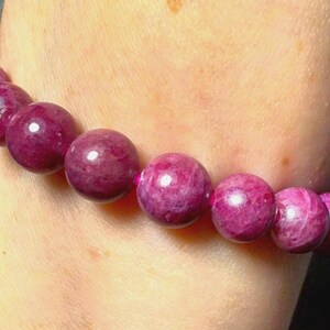 May include: A beaded bracelet featuring large, round, deep magenta beads. The beads have a polished, reflective surface and are strung together to form a bracelet. The bracelet is shown on a person's wrist.