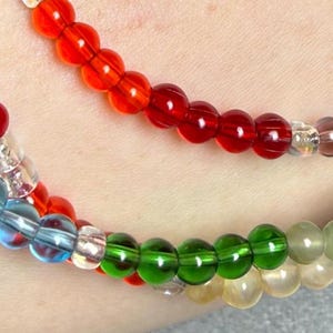 May include: Close-up of colorful beaded bracelets. The bracelets feature a variety of colors, including red, orange, blue, green, and yellow. The beads are round and appear to be made of glass or a similar material. The bracelets are worn on a wrist.