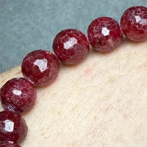 May include: A string of faceted, deep red, spherical beads arranged in a curved line. Each bead features multiple flat, polished surfaces that reflect light, creating a textured appearance across the dark crimson, semi-translucent gemstone surfaces.