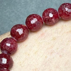 May include: A bracelet composed of round, faceted, deep red gemstone beads. Each bead features a polished, multi-faceted surface that reflects light, creating a textured appearance along the string of dark crimson stones.