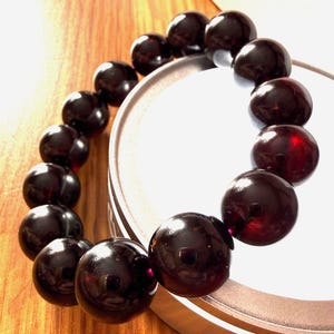 May include: A bracelet composed of smooth, spherical, dark reddish-brown beads. The beads have a polished, reflective surface that catches light, revealing deep crimson tones within the dark material. The beads are strung together in a circular loop.