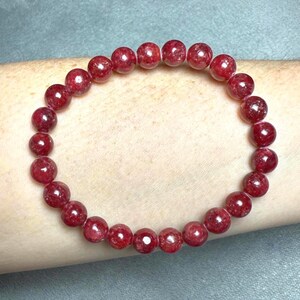 May include: A circular stretch bracelet composed of numerous small, polished, deep red spherical beads. The beads are uniform in size and shape, featuring a glossy, reflective surface that highlights their rich, dark crimson color throughout the piece.