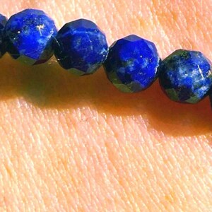 May include: A bracelet composed of multiple deep blue, faceted lapis lazuli beads. Each bead features a polished, multi-sided surface that reflects light, creating a textured appearance along the circular strand of the jewelry piece.