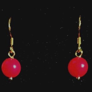 May include: A pair of gold-colored earrings with a red, round bead. The earrings have a simple hook design and a small, gold-colored bead at the bottom of the red bead. The background is black.