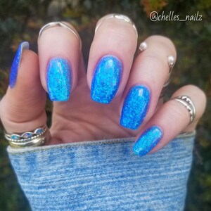 May include: Close-up of a hand with five fingernails painted in a bright blue glitter nail polish. The nails are shaped into a square with rounded edges. The hand is wearing several silver rings.