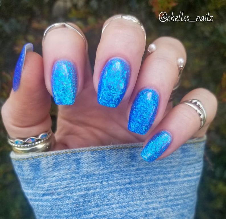 May include: Close-up of a hand with five fingernails painted in a bright blue glitter nail polish. The nails are shaped into a square with rounded edges. The hand is wearing several silver rings.