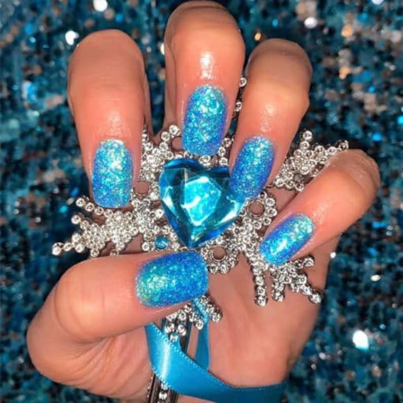 May include: Close-up of a hand with blue glitter nail polish. The nails are decorated with a snowflake design and a blue gem.