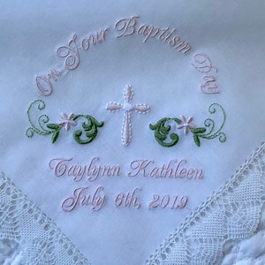 May include: White embroidered handkerchief with pink and green floral designs, a white cross, and the text "On Your Baptism Day Taylynn Kathleen July 6th, 2019".
