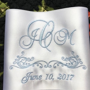 May include: White satin ribbon with light blue embroidered initials "A C M", decorative scrollwork, and the date "June 10, 2017". The ribbon is likely for a wedding or special event.