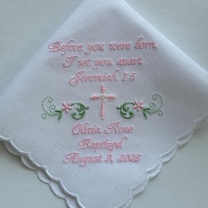 May include: White handkerchief with scalloped edges, embroidered with pink text and a cross. The text reads "Before you were born, I set you apart Jeremiah 1:5 Olivia Rose Baptized August 9, 2025". Floral and leaf accents.