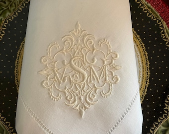 May include: A white linen napkin with a white embroidered monogram in a decorative floral design.