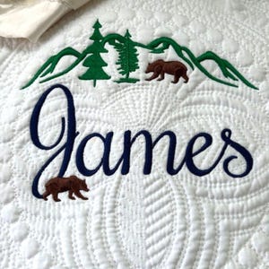 May include: White quilted blanket with embroidered forest scene and the name "James" in navy blue script. The design includes green mountains, trees, and brown bears. The blanket has a cream-colored border.