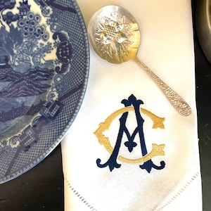 May include: White linen napkin with embroidered gold and navy blue monogram. A silver serving spoon with a floral design rests on the napkin. A blue and white patterned plate is partially visible.