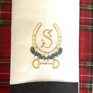 May include: White hand towel with a black border. The towel features an embroidered gold horseshoe with a green wreath and a gold letter "S" inside the horseshoe. The horseshoe is topped with a gold stirrup.