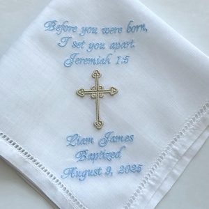Könnte beinhalten: Ein weißes Taschentuch mit Hohlsaum und hellblauer Stickerei mit dem Text Before you were born, I set you apart, Jeremiah 1:5, Liam James, Baptized, August 9, 2025, sowie einem mittig platzierten, goldfarbenen gestickten Kreuz.