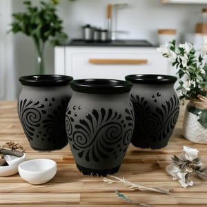 Op de afbeelding: Drie grijze keramische vazen met zwarte randen en bodems. Elke vaas heeft een zwart, wervelend bloemmotief. De vazen zijn gerangschikt op een houten oppervlak, met een witte kom en decoratieve elementen op de voorgrond.